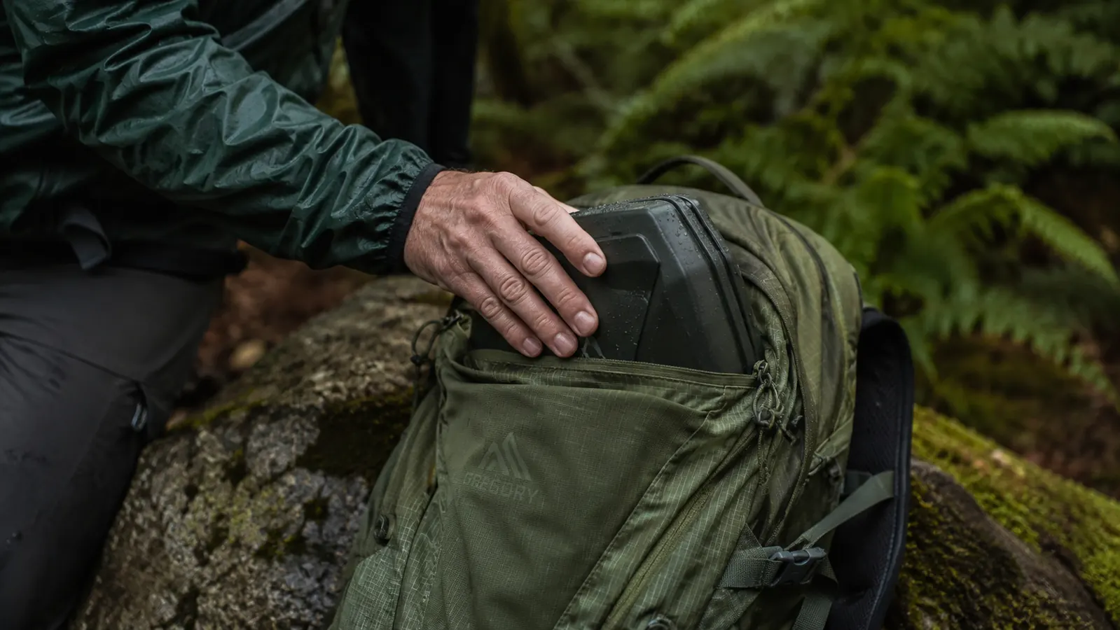 A hand tucking a folded Marmot Precip Eco rain jacket into the top pocket of a hiking daypack