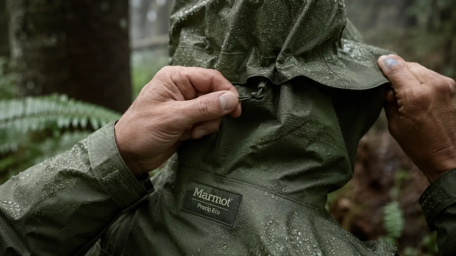 Close-up of hands cinching the hood drawcord and stiff brim of a green Marmot Precip Eco hardshell jacket, rain droplets on the fabric.