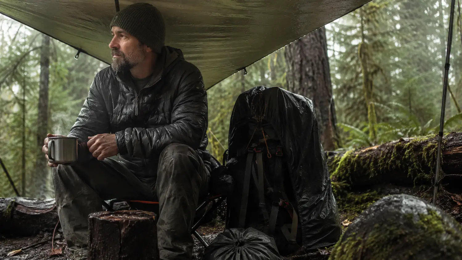 A rain-swept camp in the Cascades with steam rising from a camp mug and a pack leaned against a tree under a tarp
